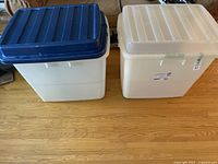 Two large storage containers side by side on a wooden floor, one with blue lid, one with white lid