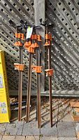 Four long adjustable metal clamps with orange clamp heads leaning against a wooden lattice fence outdoors.