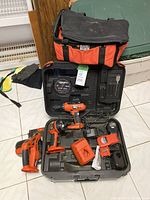 Black & Decker cordless tools including drill, saw, and impact driver arranged in a hard plastic case with charger and battery, plus a soft carrying bag beside them.