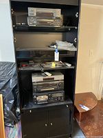 Black shelving unit containing multiple electronic audio/video components and cords, with papers on middle shelves and nearby small brown table.
