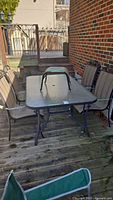 Photo of rectangular glass-top patio table with four metal and mesh fabric chairs around it, placed on wooden deck near brick wall and wooden stairs.