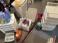 Photo showing assorted white and clear plastic storage drawer units, white laundry basket, and cleaning supplies including detergents and brushes on carpet floor.