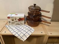 Photo showing boxed Stokes Table lobster set, three stacked brown glass Visions cookware pots with handles, and blue/white checked dish towel