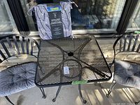 Photo of black metal mesh square table with umbrella hole and two black metal chairs with blue paisley cushions. Includes two extra chair pads with black and white leaf pattern on top of table.