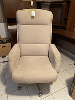 Front view of beige fabric office chair showing padded seat, backrest, and armrests with caster wheels beneath.