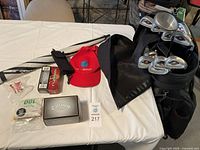 Photo shows golf accessories on table including Callaway golf balls box, Maxfli tees pack, red cap, and smaller golf items laid next to golf bag.