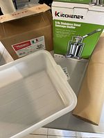 Photo showing the Reber stainless steel sausage stuffer original box, Kitchenner sausage stuffer box, white plastic meat tub, and roll of brown sausage wrapping paper.