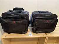 Two black Swiss Gear travel bags standing side by side on wooden surface, showing front and side views with logo visible
