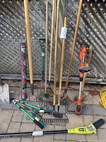 Photo showing all garden tools arranged against a lattice wooden fence including rakes, hoes, shears, digging fork, and electric hedge trimmer on ground