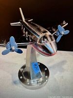 View of stainless steel airplane light on curved metal stand, showing front and part of side with blue propellers and cockpit detail