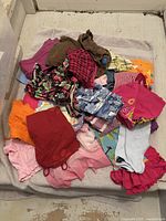 Mixed girls clothing items in a pile showing a variety of colors including red, pink, orange, brown, purple, and patterns like plaid and floral.