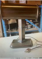 Full front view of vintage copper coloured metal desk lamp with rectangular stepped base and cylindrical shade; lamp cord and plug visible on surface.