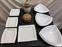 Overview of 9-piece white porcelain dish set displayed on black table featuring 3 triangular bowls, 3 rectangular plates, 1 square plate, teapot, and covered pot.