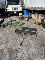 Clear front view of the metal lawn aerator and another garden tool in the background, showing the handle, tine assembly, and wheels on an asphalt surface with trucks nearby.