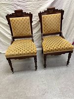 Pair of antique wooden chairs with mustard-yellow patterned upholstery, carved crest rails, turned legs with casters, against a white fabric background.