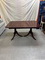 Front view of the antique mahogany wood dining table with the leaf inserted, showing the table's rectangular surface and double pedestal base with curved legs ending in brass feet caps.