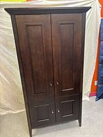 Front view of tall brown wooden armoire with two large upper paneled doors and two smaller lower paneled doors, all closed, standing on four legs with beveled top edge.