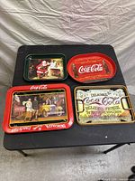 Four vintage Coca-Cola metal serving trays arranged on a black table showing front designs