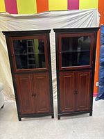 Full view of both narrow wooden corner cabinets side by side with two-tone reddish-brown and black finish, glass upper doors, and solid lower doors