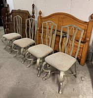 Four decorative metal chairs positioned in a row. The chairs have beige fabric seats, metal backs with decorative twisted designs, and curved scroll-style metal legs. Wood furniture is in the background.