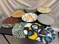 Multiple decorative plates and glass serving dishes arranged on black table, showing diverse colorful patterns including abstract, animal print, leaf motif, and solid gold colors.