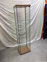 Full view of the tall narrow glass display cabinet showing wooden top and base with clear glass sides and shelves inside.
