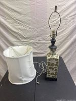 Table lamp with its ceramic base decorated with green floral pattern on white, metal neck and base accents, sitting on a black surface against a white fabric backdrop. The white fabric lampshade is placed beside the lamp and shows minor knicks and wear.
