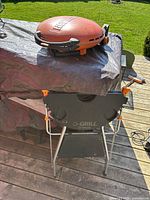 Closed view of the round orange-red Napoleon portable propane gas grill on foldable black stand on wooden deck outdoors.