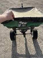 Front view showing green plastic hopper with Rugid label, held by hand to illustrate size and condition.