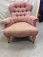 Front-facing view of vintage pink floral armchair showing button tufted backrest, plush seat, rolled armrests, wooden turned legs with casters.