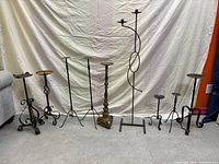 Full view of all 9 rustic metal candle holders arranged in a row on floor against white fabric background. Varied designs and heights visible, with some showing visible rust and wear.