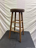 Full side view of the rustic wood bar stool with brown faux leather round seat showing wooden legs with scuffs and footrest rungs.