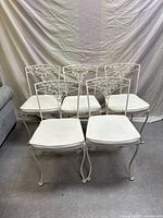 Front view of five off-white iron dining chairs with scrollwork backs and white seat cushions arranged in a row.