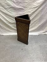 Front angled view showing the triangular shape, woven wicker sides with natural brown tone and dark wooden trim on top and bottom edges. Lightly used condition is visible.