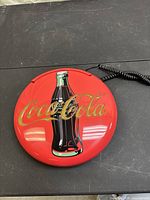 Round red Coca-Cola branded wall phone with coke bottle graphic on the front lying flat on a black surface, showing logo and color details.