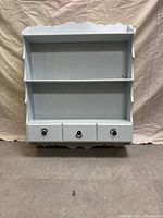 Front view of the wooden wall shelf painted light blue, showing three shelves and three drawers with metal ring pulls below.