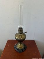 Clear view of the antique brass oil lamp with intact clear glass chimney and ornate cast brass base sitting on red surface.