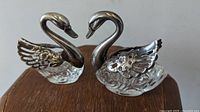 Pair of swan-shaped salt cellars with silver-tone metal necks and clear glass bodies, shown on wooden surface.