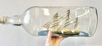 Side view of the large glass bottle showing the detailed three-masted ship model inside, resting on a blue base; clear glass with cork stopper.