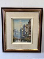 Framed oil painting showing a street scene in Paris with buildings and pedestrians, mounted with fabric mat and dark wood frame.