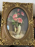 Close-up frontal photo of the framed floral print showing pink roses in a vase under bowed glass with ornate gold-brown frame.