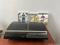 Front view of the Sony PlayStation 3 console with three game cases placed behind it: Call of Duty: World at War, FIFA 12, and FIFA 15.
