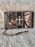 Front view of the zippered women's wallet showing the camouflage design, black vertical stripes with white stitching, and the silver-tone cross emblem with pearl accents.