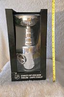 Front view of the boxed Stanley Cup shaped coin bank with measuring tape next to it showing height.