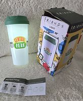 Photo of white coffee cup shaped light with green lid and Central Perk logo printed on the front, alongside original colorful box and instruction sheet.