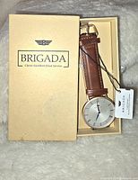 New men's Brigada watch in original beige box, showing round silver dial and brown leather strap with tag attached.