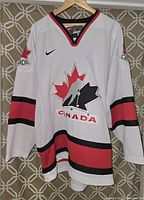 Full front view of white Nike Team Canada hockey jersey with red and black striping and maple leaf hockey logo.