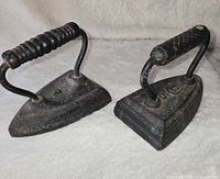 Photo showing two cast iron sad irons, one triangular base and one rectangular base, both with black wooden handles on light background.