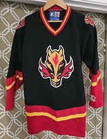 Front view of black Calgary Flames youth hockey jersey with flame horse logo centered, red and yellow accents on collar and sleeves, hanging on wooden hanger against geometric patterned background.