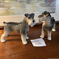 Pair of ceramic schnauzer dog figurines shown from front and side angles on a wooden surface in front of a painting.